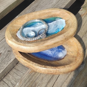 Puede incluir: Dos bandejas ovaladas de madera con un diseño de resina azul y blanca. Una bandeja contiene una pulsera de plata y dos anillos. Las bandejas son de madera marrón claro y son adecuadas para joyas o objetos pequeños.