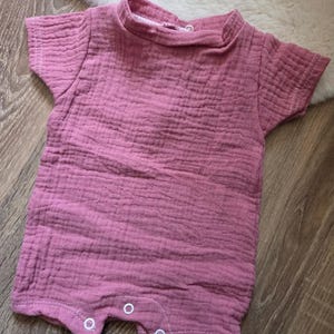Peut inclure: Un barboteuse pour bébé rose poussiéreux en tissu texturé. La barboteuse à manches courtes a une encolure ronde et des fermetures à boutons-pression en bas. Le vêtement est posé sur une surface en bois.