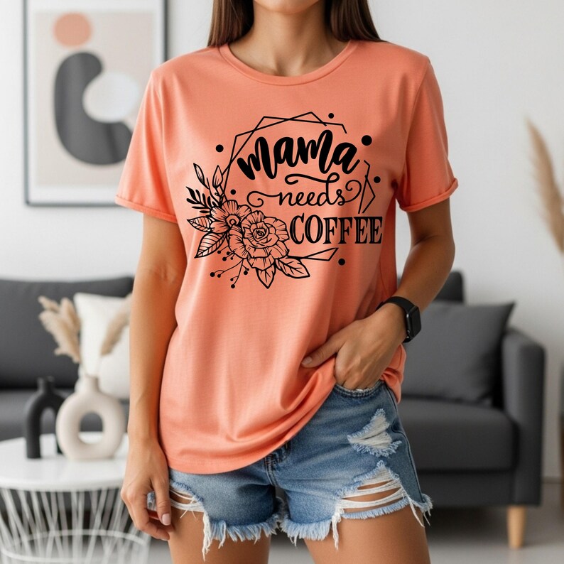 May include: Peach-colored t-shirt with the text "Mama needs COFFEE" in black script and a floral design. The shirt has short sleeves and a crew neck. The model is wearing denim shorts and a black watch.