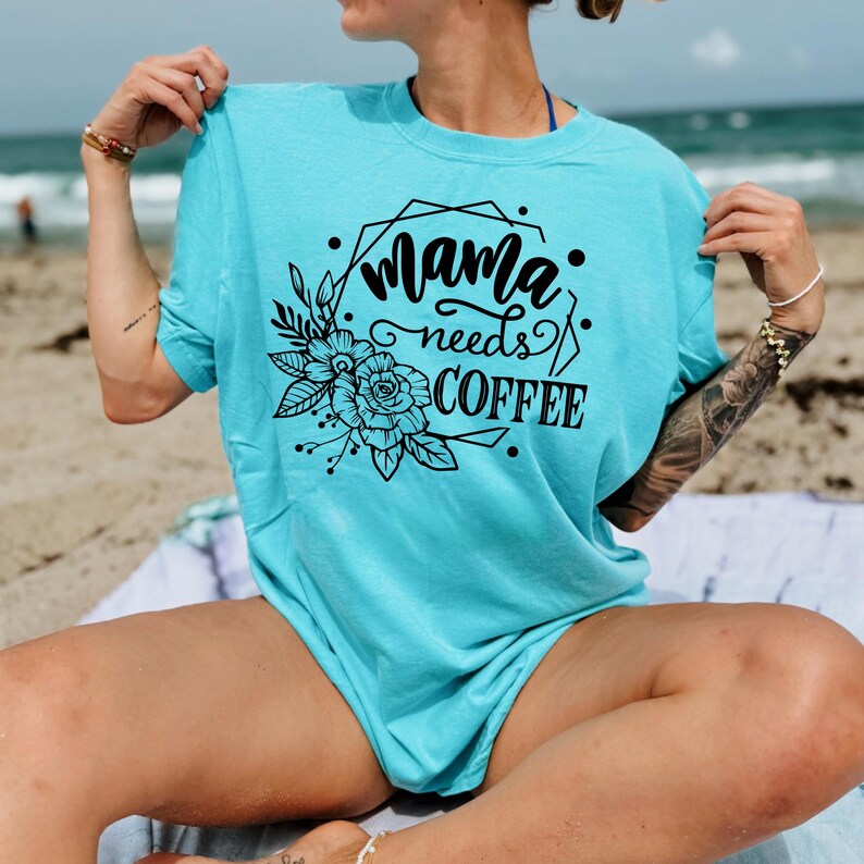 May include: A turquoise t-shirt with the text "Mama needs COFFEE" in black script and a floral design. The shirt is a short-sleeved, crew neck style. The image is taken outdoors, with a beach and ocean in the background.