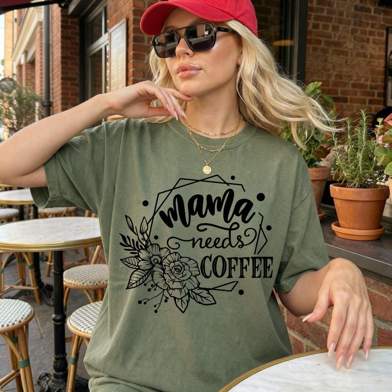May include: Olive green t-shirt with the text "Mama needs COFFEE" in black, surrounded by floral and geometric designs. The shirt is a casual style, suitable for everyday wear.