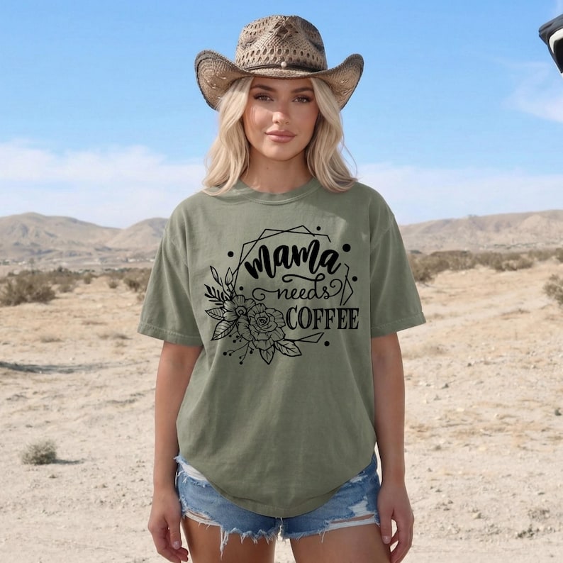 May include: An olive green t-shirt with the text "Mama needs Coffee" in a black floral design. The shirt is worn with denim shorts and a straw cowboy hat. The background shows a desert landscape.