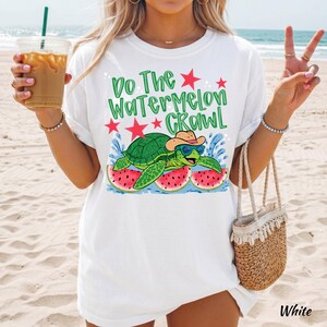 May include: A white t-shirt featuring a graphic of a green turtle wearing a cowboy hat and blue sunglasses, resting on watermelon slices. The text reads "Do The Watermelon Crawl" in green letters, accented by red stars and blue water splashes.