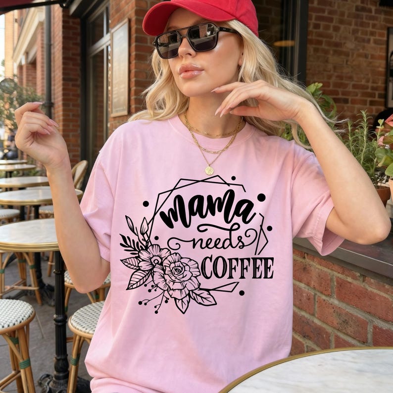 May include: A light pink t-shirt with the text "Mama needs coffee" in black script and a floral design. The shirt is a casual, oversized fit.