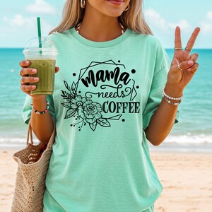 May include: A light green t-shirt with the text "Mama needs COFFEE" in black script and a floral design. The shirt is worn on a beach with a drink in hand.