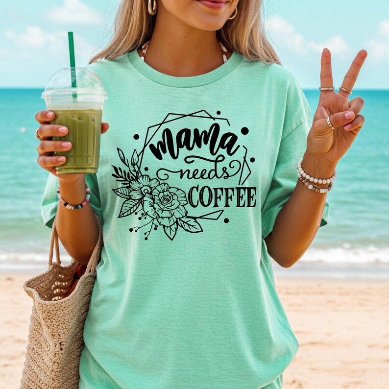 May include: A light green t-shirt with the text "Mama needs COFFEE" in black script and a floral design. The shirt is worn on a beach with a drink in hand.