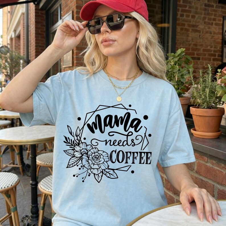 May include: Light blue t-shirt with the text "Mama needs COFFEE" in a black, decorative font. The design includes floral and geometric elements. The shirt is a casual, everyday wear item.