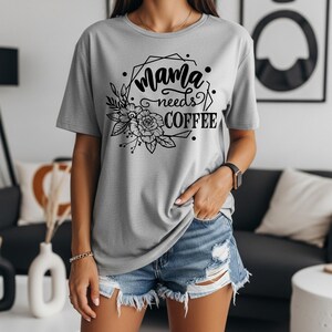 May include: A light gray t-shirt with the text "Mama needs coffee" in a black floral design. The design includes flowers, leaves, and geometric shapes. The shirt is short-sleeved and made of a soft material.