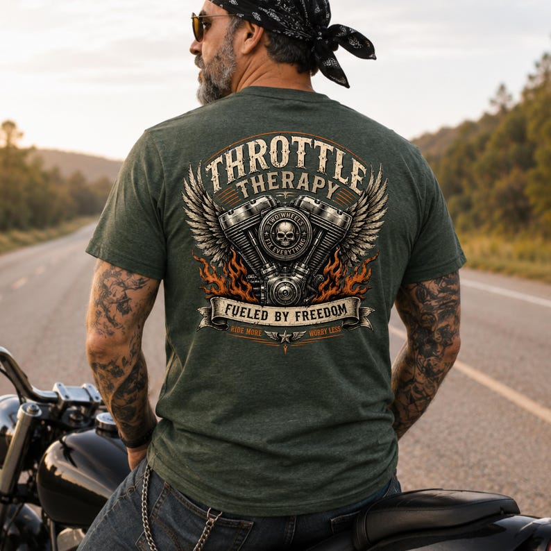 May include: A heathered olive green t-shirt featuring a large back graphic of a motorcycle engine with wings, flames, and a skull. The text reads THROTTLE THERAPY, FUELED BY FREEDOM, TWO WHEELS FIX EVERYTHING, RIDE MORE, and WORRY LESS.
