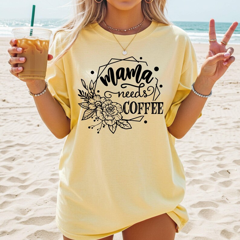 May include: A light yellow t-shirt with the text "Mama needs COFFEE" in a black floral design. The shirt is a loose fit and is being worn on a beach.