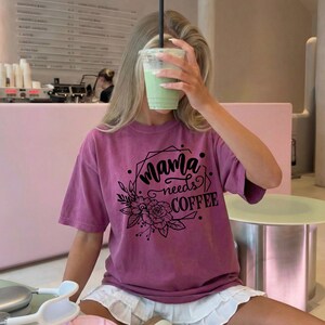 May include: A mauve-colored t-shirt with the text "Mama needs COFFEE" in a black, decorative font. The design includes floral and geometric elements. The person is holding a green drink in a clear plastic cup with a black straw.