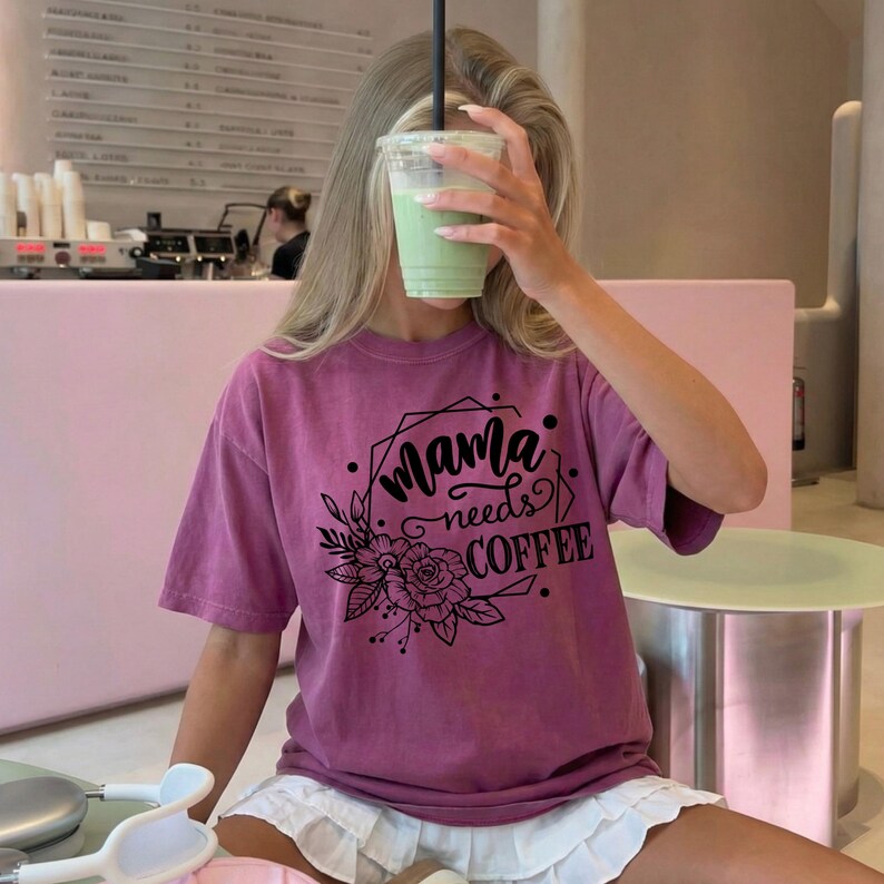 May include: A mauve-colored t-shirt with the text "Mama needs COFFEE" in a black, decorative font. The design includes floral and geometric elements. The person is holding a green drink in a clear plastic cup with a black straw.
