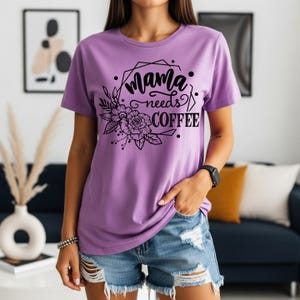 May include: A lilac-colored t-shirt with the text "Mama needs COFFEE" in a black floral design. The shirt is short-sleeved and has a round neckline. The design includes a geometric frame and floral elements.
