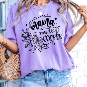 May include: A lilac-colored t-shirt with the text "Mama needs COFFEE" in black script and block letters, surrounded by a floral and geometric design. The shirt is a casual, short-sleeved style.