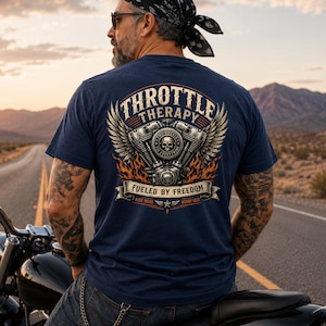 May include: A navy blue t-shirt featuring a graphic on the back with a motorcycle engine, wings, and flames. The text reads "THROTTLE THERAPY," "FUELED BY FREEDOM," "TWO WHEELS FIX EVERYTHING," and "RIDE MORE WORRY LESS" in a stylized, vintage-inspired font.