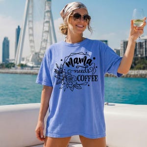 May include: A light blue t-shirt with the text "Mama needs COFFEE" in black script and a floral design. The shirt is worn by a person wearing sunglasses and a headscarf, holding a glass of wine.