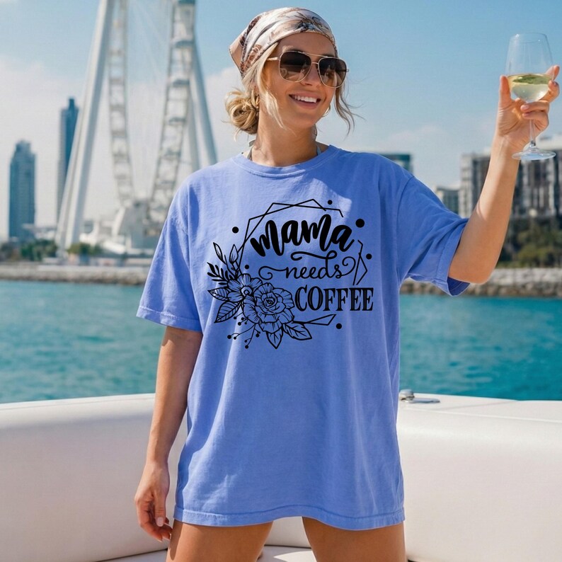 May include: A light blue t-shirt with the text "Mama needs COFFEE" in black script and a floral design. The shirt is worn by a person wearing sunglasses and a headscarf, holding a glass of wine.