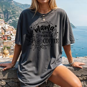 May include: A charcoal gray t-shirt with the text "Mama needs COFFEE" in a black floral design. The shirt is a relaxed fit and has short sleeves. The design includes a floral arrangement and geometric shapes.