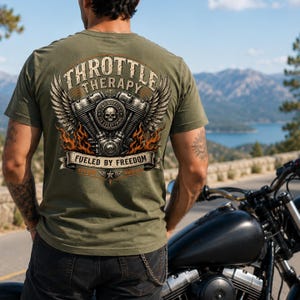 May include: An olive green t-shirt featuring a large back graphic of a motorcycle engine with wings, flames, and a skull. The text reads "THROTTLE THERAPY", "TWO WHEELS FIX EVERYTHING", and "FUELED BY FREEDOM" in a vintage, distressed font style.