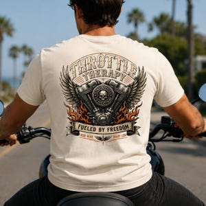 May include: A cream-colored t-shirt featuring a back graphic of a motorcycle engine with wings, a skull, and flames. The text reads THROTTLE THERAPY, FUELED BY FREEDOM, TWO WHEELS FIX EVERYTHING, and RIDE MORE WORRY LESS in a detailed, vintage-style design.