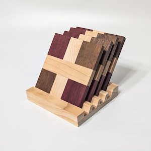 May include: A set of six wooden coasters with a geometric design, featuring alternating squares of light, medium, and dark wood tones. The coasters are displayed in a matching wooden holder, showcasing a natural wood grain.