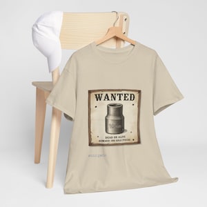 May include: A beige t-shirt featuring a graphic of a 10mm socket on a wooden-framed wanted poster. The text reads WANTED, 10mm, DEAD OR ALIVE, REWARD: 500 GOLD PIECES, and gear gear. A white baseball cap hangs on the wooden chair behind the t-shirt.