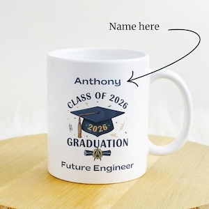 Peut inclure: Mug en céramique blanche sur le thème de la remise de diplômes. La tasse présente le nom "Anthony", le texte "Class of 2026 Graduation" et "Future Engineer". Une toque et un parchemin bleus sont également affichés.