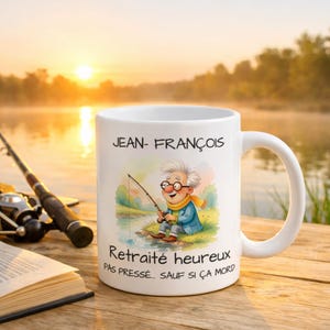 Puede incluir: Taza de cerámica blanca con una ilustración de dibujos animados de un hombre pescando. La taza tiene el nombre "JEAN-FRANÇOIS" y el texto "Retraité heureux PAS PRESSÉ... SAUF SI ÇA MORD". Una caña de pescar y un libro abierto están en el fondo.