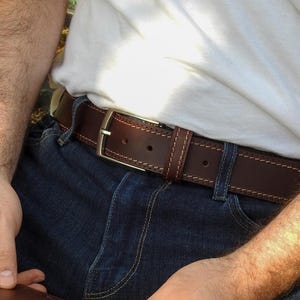 Puede incluir: Un cinturón de cuero marrón con hebilla plateada se lleva sobre unos vaqueros azules. El cinturón tiene un diseño cosido y se lleva con una camiseta blanca. El cinturón mide aproximadamente 3,8 cm de ancho.