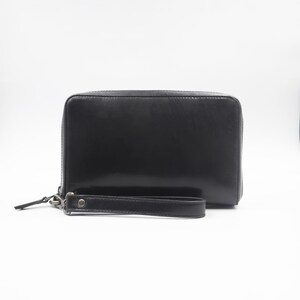 Puede incluir: Un monedero de pulsera de cuero negro con cierre de cremallera. La cartera tiene una correa de muñeca sujeta con un cierre de metal plateado. El monedero es rectangular y parece estar hecho de cuero liso.