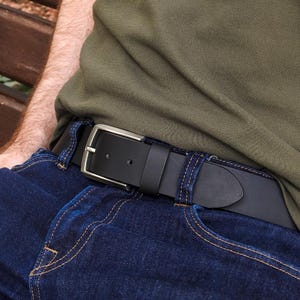 Puede incluir: Un cinturón de cuero negro con hebilla plateada abrochado sobre vaqueros azules. El cinturón se lleva con una camisa verde. La hebilla es rectangular y el cinturón mide unos 3,8 cm de ancho.