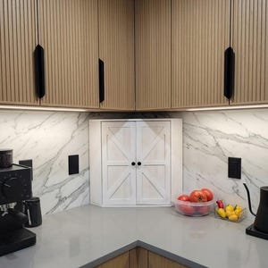 Puede incluir: Una esquina de cocina moderna con armarios de madera clara y tiradores negros, complementada con una encimera de mármol blanco. Un armario blanco con pomos negros está centrado debajo. Tomates y pimientos frescos sobre la encimera.