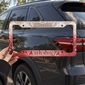 May include: A rectangular license plate frame featuring a gradient design that transitions from a beige top with clouds, wings, and a halo to a red bottom with flames and a devil tail. The text DRIVE CUTE is at the top and STAY WILD is at the bottom.