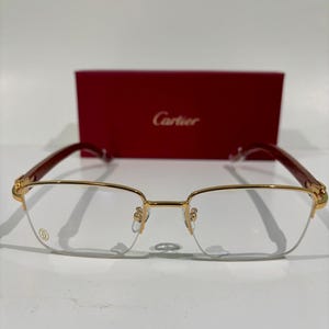 Puede incluir: Gafas con montura dorada de forma rectangular y varillas marrón rojizo. Las gafas están colocadas delante de una caja roja con el nombre de la marca "Cartier" en letras doradas. Las lentes son claras.