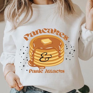 Peut inclure: Sweat-shirt blanc avec l'inscription "Pancakes & Panic Attacks" en lettres orange. Le motif représente une pile de crêpes avec du sirop et une noix de beurre. Des éclats d'étoiles noirs sont dispersés autour du motif.