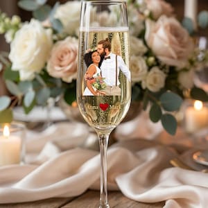 Puede incluir: Una copa de champán llena de líquido burbujeante, con una imagen personalizada de una pareja. La copa está colocada sobre un fondo de flores y velas, con los nombres "Anna" y "Mark" y un símbolo de corazón impreso en el cristal.