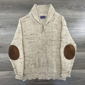 May include: A cream-colored, zip-up sweater with a shawl collar and brown elbow patches. The sweater has two front pockets and a small label near the collar. The zipper has a circular pull. The sweater is made of a thick, textured knit.