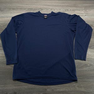 May include: A solid navy blue long-sleeve shirt with a crew neckline and a small rectangular brand label located at the inside of the collar. The garment features a smooth, uniform texture and long sleeves with finished cuffs at the wrists.