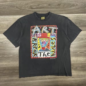 May include: Black t-shirt with a colorful graphic that reads "ART BREAK RULES The." The graphic features a stylized face with a red hat and glasses. The shirt has a crew neck and short sleeves.