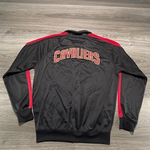 May include: A black track jacket featuring red stripes running down the length of each sleeve. The back of the jacket displays the word CAVALIERS in large, arched, gold-colored lettering with a red outline, centered across the upper back area.