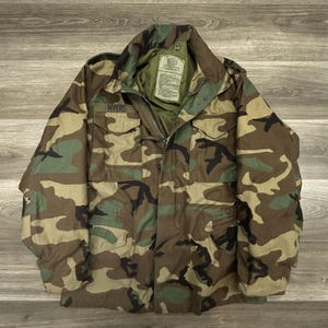 Pode incluir: Um casaco de campo com padrão de camuflagem florestal em tons de castanho, verde e preto. O casaco possui gola alta, fecho de correr frontal com aba de botões de pressão, dois bolsos no peito e o nome MYERS impresso acima do bolso.