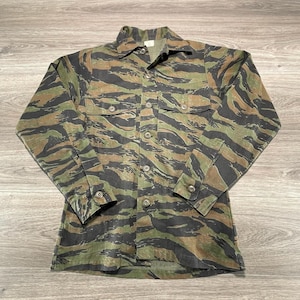 Puede incluir: Una camisa de manga larga con botones y un patrón de camuflaje de rayas de tigre en tonos verde oliva, marrón y negro. La camisa incluye un cuello puntiagudo, dos bolsillos en el pecho con solapas abotonadas y puños abotonados en cada manga.