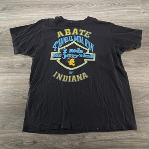 May include: A black short-sleeve t-shirt featuring a graphic with the text ABATE 4th ANNUAL MDA RUN, I RODE FOR JERRY'S KIDS, and OF INDIANA. The design includes a yellow and light blue emblem with a small yellow cartoon face illustration.