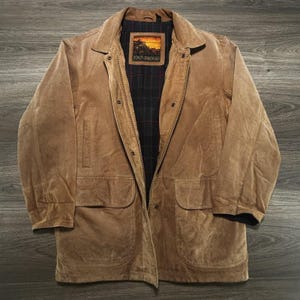 May include: A tan suede jacket featuring a full-length front zip and snap button fastening. The interior is lined with a black fabric featuring a red plaid pattern and a rectangular patch label that reads Out Brook with a sunset landscape graphic.