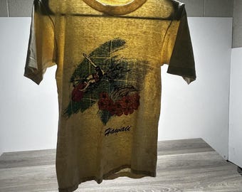 May include: A pale yellow short-sleeve t-shirt featuring a graphic print of a person surfing on a wave, accompanied by red hibiscus flowers and the word Hawaii printed in black script lettering below the central illustration on the front.