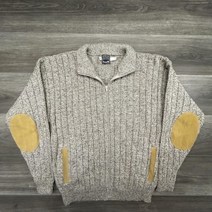 May include: A beige and grey marled knit zip-up sweater featuring a ribbed texture, a high collar, and long sleeves. The garment includes tan suede-like elbow patches and matching vertical trim accents on the two front pockets with a central zipper.