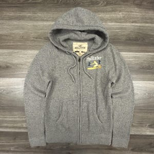 May include: A gray zip-up hoodie with a hood and front pockets. The hoodie features the "Hollister" logo on the chest and a small graphic of a surfer on a surfboard. The jacket has a full zipper and drawstrings.