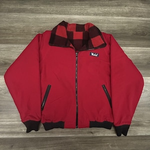 May include: A red zip-up jacket featuring a black and red buffalo plaid collar, black ribbed cuffs and hem, and two slanted zippered pockets. A small rectangular blue label with the white text Woolrich is stitched on the upper left chest area.