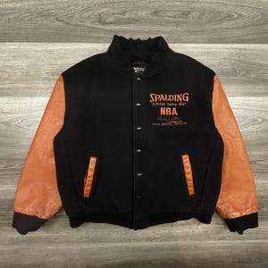May include: Black and orange Spalding NBA varsity jacket. The jacket has black fabric with orange leather sleeves and pockets. The text "SPALDING Official Game Ball NBA" is printed in orange on the chest.