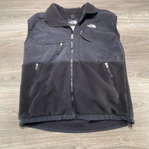 May include: A black sleeveless vest featuring a smooth upper panel and a textured fleece lower panel. It includes a full-length front zip, a high collar, two zipped hand pockets, and a zipped chest pocket with The North Face logo branding.